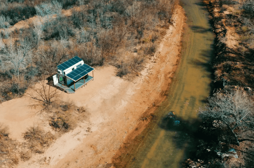 Aerial view of a small house with solar panels beside a muddy river, surrounded by dry, barren land and sparse trees.